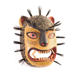 May include: Hand-painted wooden mask depicting a fierce jaguar. The mask features a yellow and black spotted pattern, red eyes, and a snarling mouth with visible teeth. Black hair-like accents surround the face, adding to its dramatic appearance.