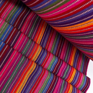 Puede incluir: Una tela tejida con un vibrante patrón de rayas multicolores. El textil muestra líneas verticales repetitivas en tonos magenta, morado, naranja, verde y azul, acentuadas por finas rayas blancas a lo largo de todo el material.