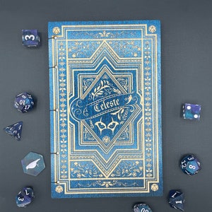May include: A blue and gold journal with the word "Celeste" on the cover, surrounded by intricate floral designs. The journal is surrounded by various blue and silver dice.