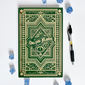 Puede incluir: Libro verde con un diseño dorado y las palabras "Dungeon Master" en la portada. El libro está rodeado de dados azules.
