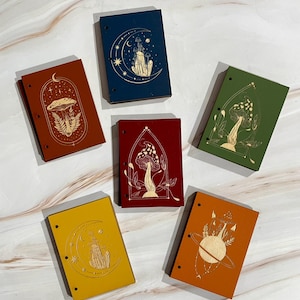 May include: A collection of six small, colorful notebooks with gold-colored celestial and mushroom designs. The notebooks are in various colors, including red, blue, green, yellow, and orange. Each notebook has a different design, such as a moon, stars, and mushrooms.