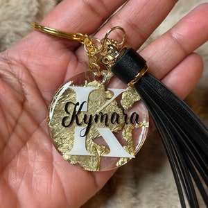 May include: Gold and white resin keyring with a black tassel. The keyring features the letter "K" and the name "Kymera" in black lettering.