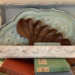 May include: A distressed white wooden coat rack with three metal hooks, featuring carved floral details. Behind it, a large wooden leaf-shaped decor and a light blue tray with embossed floral patterns. The items rest on a stack of vintage books.