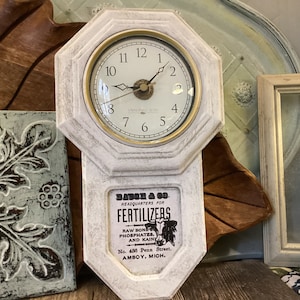 Puede incluir: Reloj octogonal blanco con una esfera dorada y agujas negras. El reloj está unido a un letrero blanco con texto negro que dice "Bauch & Co. Headquarters for Fertilizers Raw Bone Phosphates, and Kain No. 436 Penn Street, Amboy, Mich."