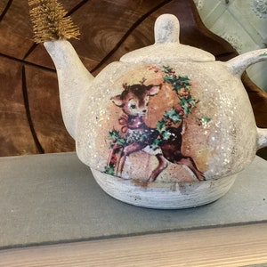 Puede incluir: Tetera de cerámica blanca con un diseño festivo que presenta un ciervo y acebo. La tetera tiene un acabado de purpurina dorada y un adorno de cepillo dorado en el pico.