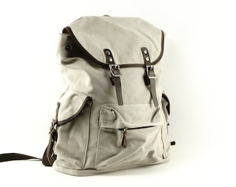 May include: A light gray canvas backpack featuring dark brown leather-like trim, two front pockets with flap closures, and a main compartment secured by a top flap with two adjustable metal buckles and dark brown straps over a zippered opening.