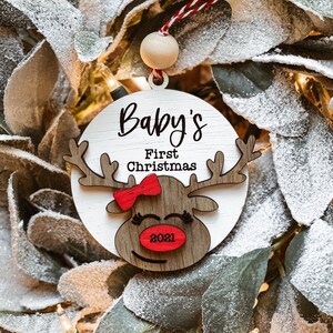 May include: White wooden ornament with a red bow and reindeer face. The ornament says "Baby's First Christmas 2021".