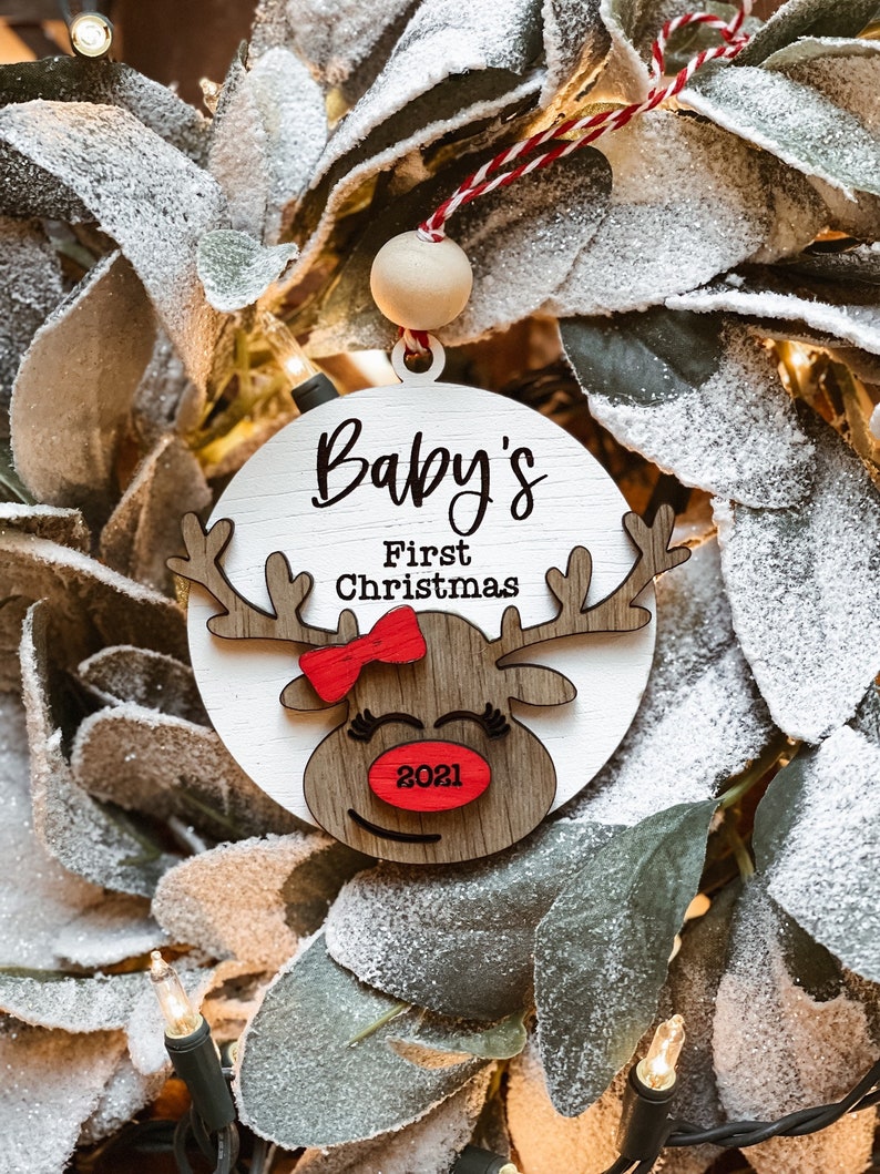 May include: White wooden ornament with a red bow and reindeer face. The ornament says "Baby's First Christmas 2021".