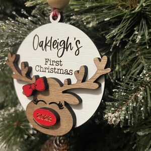May include: White wooden ornament with a reindeer design. The ornament has a red bow, a red nose with the year 2023 on it, and the text "Oakleigh's First Christmas".