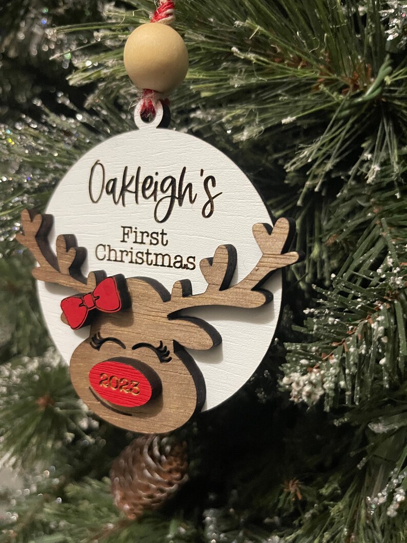 May include: White wooden ornament with a reindeer design. The ornament has a red bow, a red nose with the year 2023 on it, and the text "Oakleigh's First Christmas".