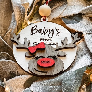 May include: A wooden reindeer ornament with a red bow and the text "Baby's First Christmas 2021".