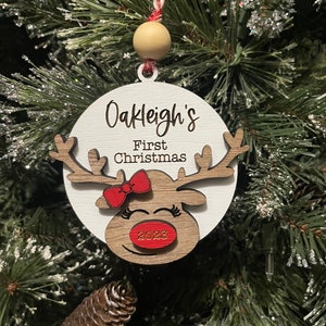 May include: White wooden ornament with a reindeer face, red bow, and the text "Oakleigh's First Christmas 2023".