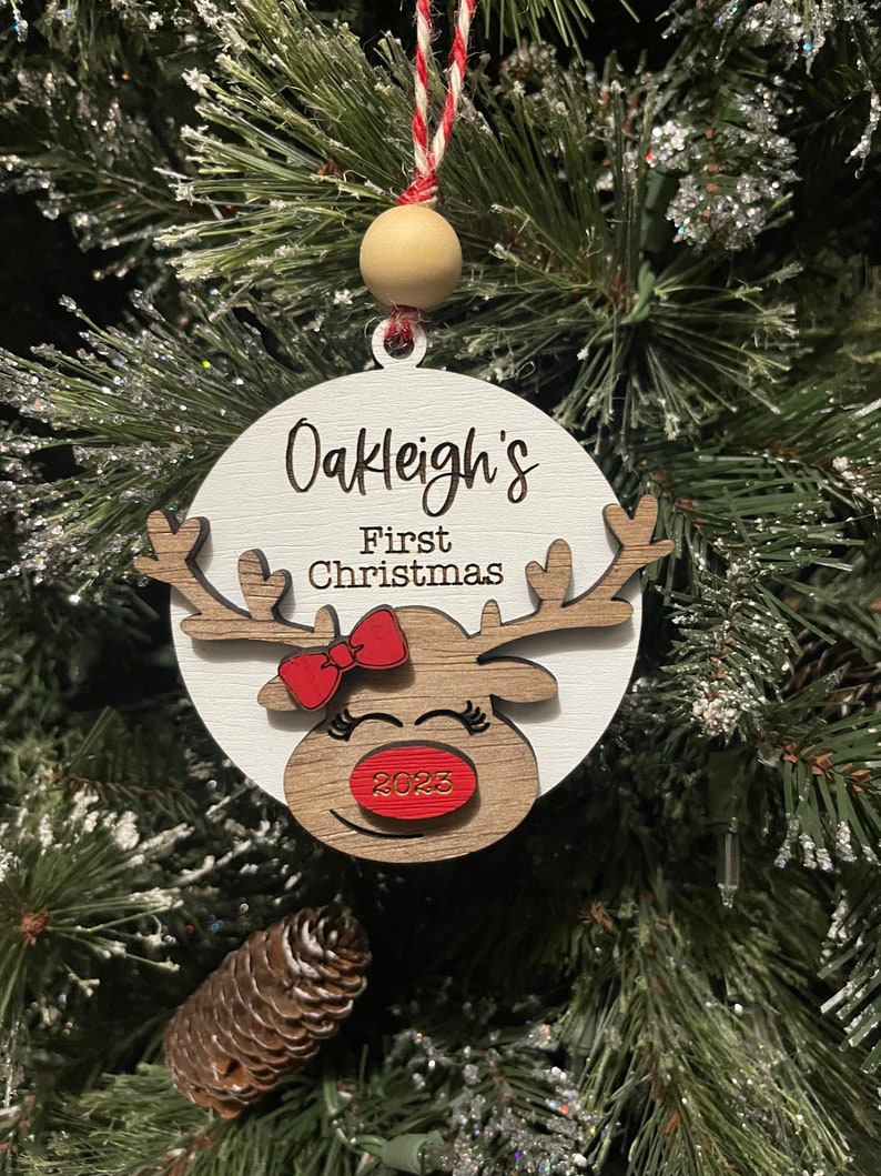 May include: White wooden ornament with a reindeer face, red bow, and the text "Oakleigh's First Christmas 2023".
