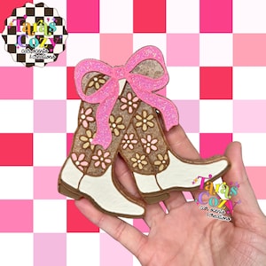 May include: A decorative car scent featuring a pair of cowboy boots with a pink bow. The boots are brown with white accents and floral designs. The background is a pink and white checkered pattern. The text "Tara's Cozy car scents & creations" is visible.
