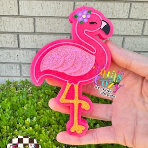 May include: A pink flamingo-shaped car air freshener with a glittery body and a flower on its head. The flamingo has gold legs and a black beak. The product is from Tara's Cozy Car Scents & Creations.
