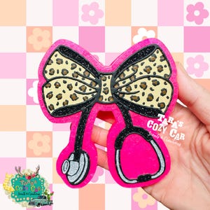May include: A pink stethoscope badge reel with a bow featuring a leopard print design. The badge reel has black glitter accents and the stethoscope heads are silver glitter. The text "Tara's Cozy Car" is visible.