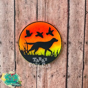 May include: A round, black car air freshener featuring a silhouette of a dog walking through tall grass with three flying birds against a sunset background. The text "Tara's Cozy Car" is at the bottom.