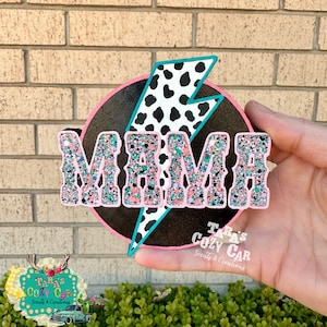 May include: A decorative sign with the word "MAMA" in glittery letters. The sign features a black background with a pink border and a cow print lightning bolt. The sign is held in a hand.