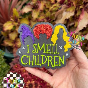 May include: A black, textured, shaped plaque featuring three glittery hair silhouettes in purple, red, and yellow. The text "I SMELL CHILDREN" is printed in bright green, accompanied by small white star accents. The design is flat and decorative.