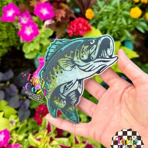 May include: A bass-shaped car scent and creation. The design features a detailed illustration of a bass fish in shades of black, green, and silver. The product is held in front of a colorful floral background. The text "Tara's Cozy Car Scents & Creations" is visible.
