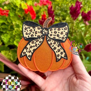 May include: An orange pumpkin-shaped air freshener featuring a prominent leopard print bow with black glitter edges and a brown stem. The text Tara's Cozy car scents & creations is visible on the right side and in a checkered logo at the bottom.