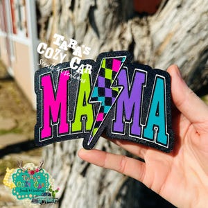 May include: A colorful "MAMA" patch with a lightning bolt design. The letters are in bright pink, green, purple, and teal, outlined in white, and set against a black denim background. The lightning bolt features a checkered pattern.