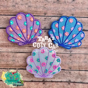 May include: Three seashell-shaped car air fresheners in purple, blue, and white, each adorned with small flower patterns in various colors. The edges are outlined with glitter. The text "Tara's Cozy Car" is visible.