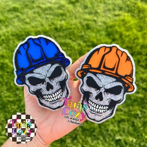 May include: Two skull-shaped car air fresheners. One has a blue hard hat, the other an orange hard hat. Both have black eyes and a menacing expression. The text "Tara's Cozy Car Scents & Creations" is visible.