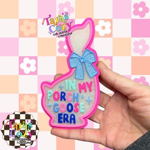 May include: A pink and white goose-shaped car air freshener with a blue glitter bow and the text "IN MY PORCH GOOSE ERA". The air freshener has a pink border and is decorated with stars.