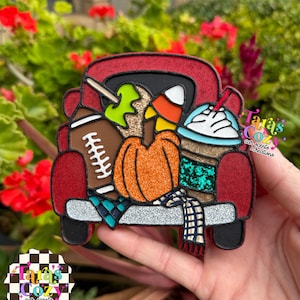 May include: A red truck-shaped ornament featuring a brown football, a green candy apple, a glittery orange pumpkin, a piece of candy corn, and a coffee cup with whipped cream. The design includes a blue plaid scarf and silver bumper accents.