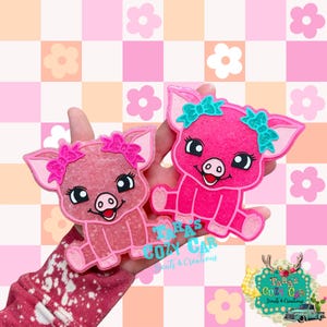 May include: Two pink pig-shaped air fresheners with bows. One pig has a pink bow, the other has a teal bow. The pigs have black eyes and smiling red mouths. The background is a pink and peach checkered pattern with flower accents.