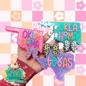 May include: Four car air fresheners in the shape of U.S. states. The Oklahoma and Texas shaped air fresheners are pink, teal, and purple with the state name and a bow. The air fresheners are decorated with glitter.