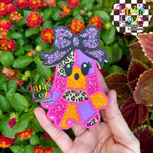 May include: A ghost-shaped ornament featuring a patchwork design of leopard print, bright orange, hot pink, and purple textures. The ghost is topped with a large, glittery purple and black bow and has two black eyes. The text Tara's Cozy car scents & creations is visible.