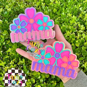 May include: Two glittery pink and purple floral designs with the word "mama" in a cursive font. The flowers have teal, orange, and purple accents. The designs are held in a hand, with a logo in the background.