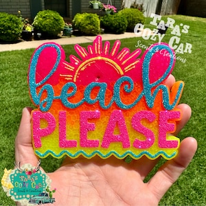 May include: A colorful, glittery car air freshener with the words "Beach Please" in bright pink and turquoise. The design includes a sun and is set against a gradient of orange and yellow. The product is held in a hand.