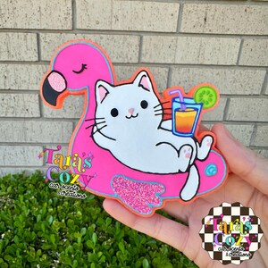 May include: A pink flamingo pool float with a white cat enjoying a drink. The cat is lounging on the float, which has a glittery pink section. A glass of orange juice with a straw and a cucumber slice are also visible.