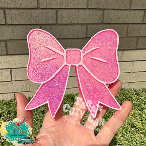 May include: A pink glittery bow-shaped car air freshener. The bow has a white outline and a square center. The text "Tara's Cozy Car Scents & Creations" is visible on the right side of the bow.