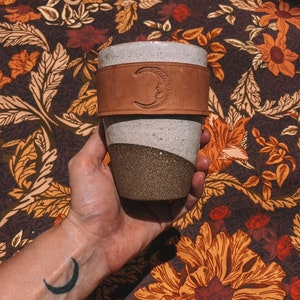 Può includere: Una tazza in ceramica bianca con una manica in pelle marrone. La manica ha un design a forma di luna crescente. La tazza è tenuta da una mano con un tatuaggio a forma di luna crescente.