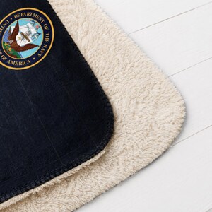 May include: Navy blue and cream-colored blanket. The blanket features the official seal of the United States Department of the Navy. The navy blue side has a ribbed texture, while the other side is a soft, cream-colored sherpa material.