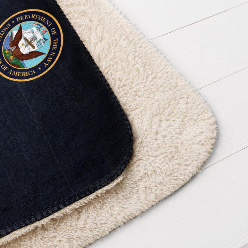 May include: Navy blue and cream-colored blanket. The blanket features the official seal of the United States Department of the Navy. The navy blue side has a ribbed texture, while the other side is a soft, cream-colored sherpa material.