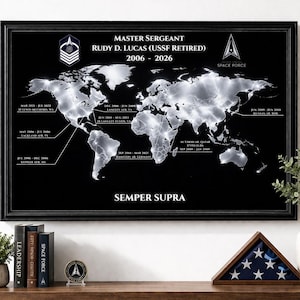 May include: A framed black and white world map with highlighted locations and text. The text reads "MASTER SERGEANT RUDY D. LUCAS (USSF RETIRED) 2006 - 2026" and "SPACE FORCE". The frame sits on a wooden surface with decorative items.