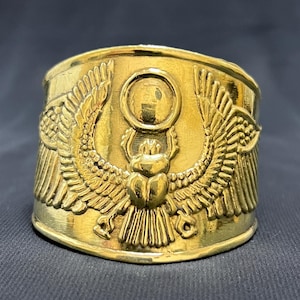 May include: A gold-colored cuff bracelet with an Egyptian scarab beetle design. The bracelet features a detailed relief of a scarab beetle with outstretched wings, a sun disc, and hieroglyphic details. The cuff is wide and has a polished finish.