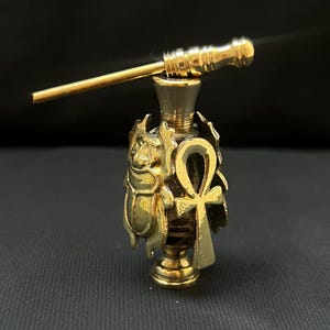 May include: A small, gold-toned metal perfume bottle featuring an Egyptian-inspired design. The bottle is adorned with a sculpted scarab beetle and an ankh symbol on its exterior. It includes a matching gold-toned applicator wand resting horizontally across the top opening of the bottle.