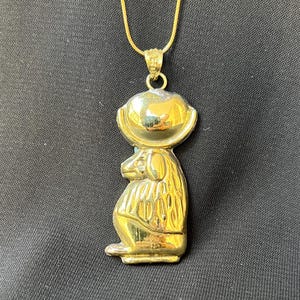 May include: A gold-colored pendant necklace featuring a meerkat design. The meerkat pendant is detailed with a textured body and a smooth, rounded head. The necklace has a delicate gold chain and a simple clasp.