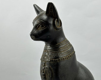 May include: Black Egyptian cat statue with hieroglyphics on the base. The cat is sitting with its head turned to the left. The statue is made of a dark, textured material.