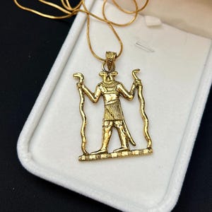 May include: A gold-toned pendant necklace featuring a cutout design of an Egyptian deity figure holding two serpents. The pendant is suspended from a thin gold-toned chain and rests inside a white, rectangular, hinged jewelry display box.