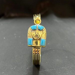 Könnte beinhalten: Ein goldfarbener Ring mit einem ägyptischen Pharao-Design. Das Gesicht des Pharaos ist goldfarben mit türkisfarbenen Akzenten. Der Ring hat ein gestapeltes Banddesign und einen kleinen, runden gelben Stein oben.