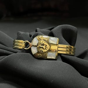 Op de afbeelding: Een goudkleurige armband met een Egyptisch geïnspireerd ontwerp. De armband heeft een centraal rechthoekig element met een gebeeldhouwd gezicht en ingelegde witte stenen. De band heeft meerdere parallelle staven. De armband wordt getoond op zwarte stof.