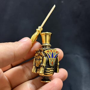 Peut inclure: Flacon de parfum doré avec un design égyptien, tenu dans une main. La bouteille présente des détails complexes, notamment des motifs de style hiéroglyphique et des accents d'émail coloré. Un applicateur doré est tenu au-dessus de la bouteille.