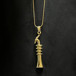 Op de afbeelding: Goudkleurige hangerketting met een gestileerd, gelaagd ontwerp en een gebogen element aan de bovenkant. De hanger hangt aan een fijne gouden ketting, tegen een donkere achtergrond. De ketting is waarschijnlijk van metaal.
